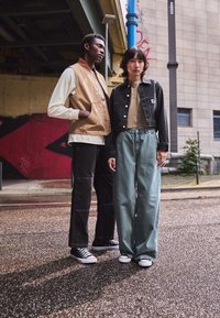 Zwei Modelle stehen auf einer nassen Oberfläche. Eines trägt eine tanfarbene Weste über einem weißen Langarmshirt, schwarze Hosen und schwarze Sneaker. Das andere trägt eine gekürzte schwarze Jacke, ein beiges Oberteil und lockere hellgrüne Hosen.