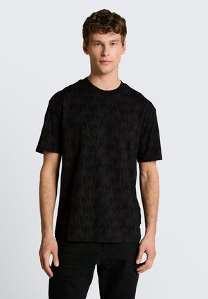 Jeune homme aux cheveux courts et bouclés portant un T-shirt noir à manches courtes avec des motifs subtils de palmiers sur un fond clair uni.