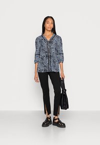 Blusa padrão preta e azul com decote em V, mangas três quartos e fendas pretas em calças pretas ajustadas. Sapatos de plataforma pretos com tiras.