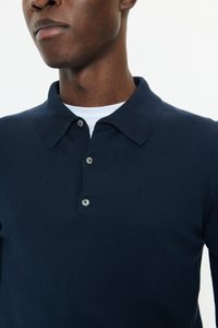 Närbild av en person som bär en marinblå långärmad pikétröja med tre knappar över en vit undertröja mot en ljus bakgrund.