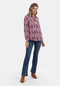 Camicia a maniche lunghe colorata con un vivace motivo geometrico nei toni del rosa, verde e blu, abbinata a jeans a zampa d'elefante in denim scuro e stivali alla caviglia marroni.