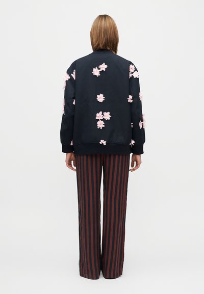 Jachetă bleumarin cu aplicații florale roz, manșete striate, asortată cu pantaloni largi, cu dungi verticale roșii și negre. Design minimalist, fără elemente metalice vizibile.