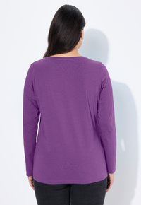 Fialové tričko s dlouhými rukávy, zaobleným výstřihem, měkkou texturou a volným střihem. Viditelné švy na lemu a manžetách.