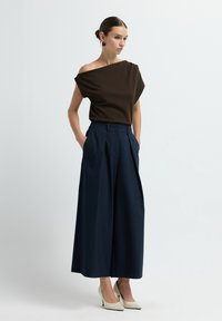 Donna che indossa un top scuro a spalle scoperte, pantaloni larghi blu navy, tacchi alti color crema e orecchini bordeaux, in piedi con le mani nelle tasche.
