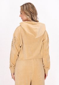 Giacca beige cropped con cappuccio in morbido tessuto, con dettagli borchiati sulle maniche. Presenta una vestibilità rilassata e polsini a coste.