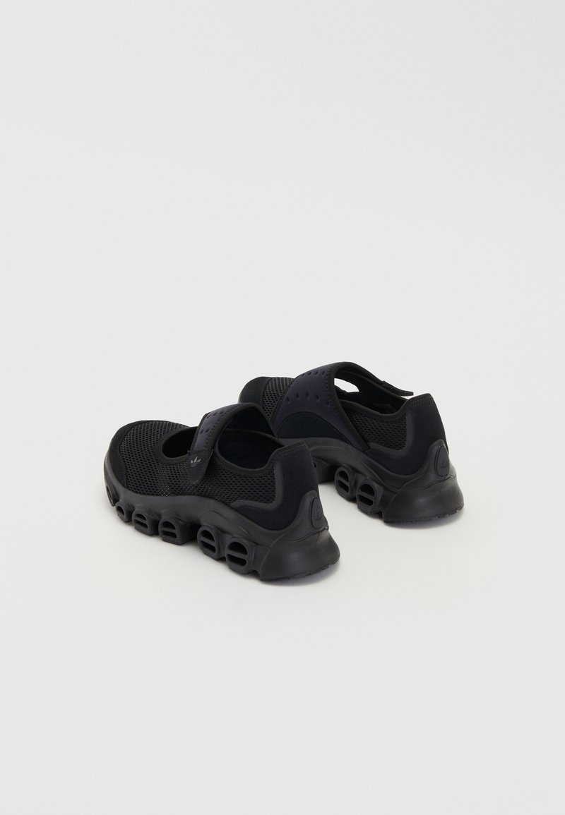 Baskets de sport noires avec une tige en maille, une sangle ajustable et une semelle texturée distinctive dotée de découpes circulaires.