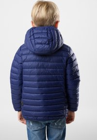 Merinottinen takki, jossa on huppu, esittelee vaakasuoria tikattu linjoja. Materiaali näyttää kiiltävältä ja kevyeltä, jota on yhdistetty sinisiin farkkuihin.