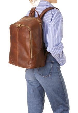 Zaino in pelle marrone con un design elegante, caratterizzato da due cerniere e cuciture a contrasto. Indossato sulla schiena, abbinato a una camicia a righe e jeans.