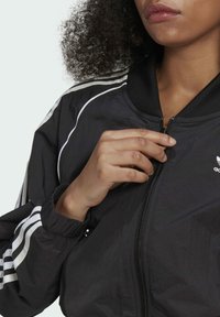 Giacca a vento nera con strisce bianche sulle spalle, chiusura con zip e polsini elasticizzati. Realizzata in materiale leggero.