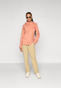 Jachetă ușoară, impermeabilă, de culoare piersică, cu un design ajustat, închidere prin fermoar și glugă ajustabilă; asortată cu pantaloni bej și sneakers neutri.