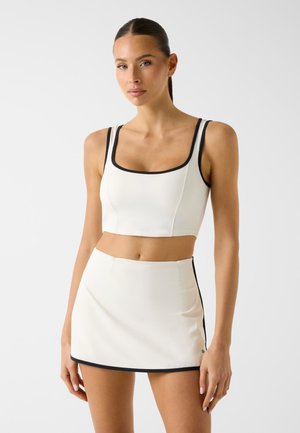 Donna che indossa un top corto bianco e una minigonna con rifiniture nere, in piedi contro uno sfondo chiaro e uniforme.