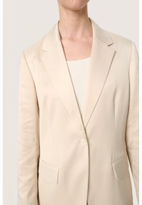 Beige színű blézer sima anyagból, kihajtott gallérral, egygombos záródással és két elülső zsebbel. Tailored fit, bélés nélküli belső.