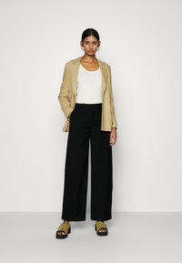 Beige blazer over een witte tanktop, gecombineerd met zwarte wijde broek en gele sandaaltjes met patroon op een platform. Eenvoudige sieradenaccenten.