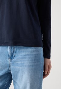 Langärmliges Marinehemd aus weichem Stoff, mit geradem Saum und einem kleinen Markenlabel an der Seite, getragen über hellblauen Jeans.