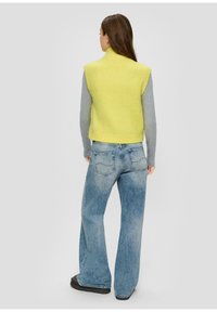 Strick-Weste ohne Ärmel in leuchtendem Gelb, über einem grauen Langarmshirt getragen, kombiniert mit hochtaillierten, hellblauen weiten Jeans.