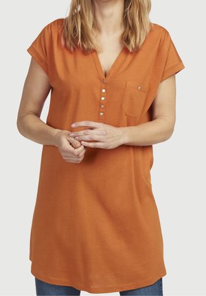 Frau trägt eine lockere orangefarbene Kurzarm-Tunika mit Metallknöpfen und Brusttasche, hält ihre verschränkten Hände vor sich.