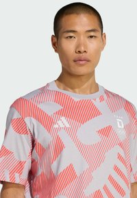 Grijs T-shirt met rood gestreept en geometrisch patroon, met wit logo en tekst. Korte mouwen, casual ontwerp, katoenachtige textuur.