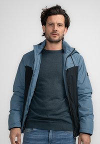 Chaleco aislante azul y negro con cierre de cremallera y capucha, que presenta una textura suave y paneles en contraste en los hombros.