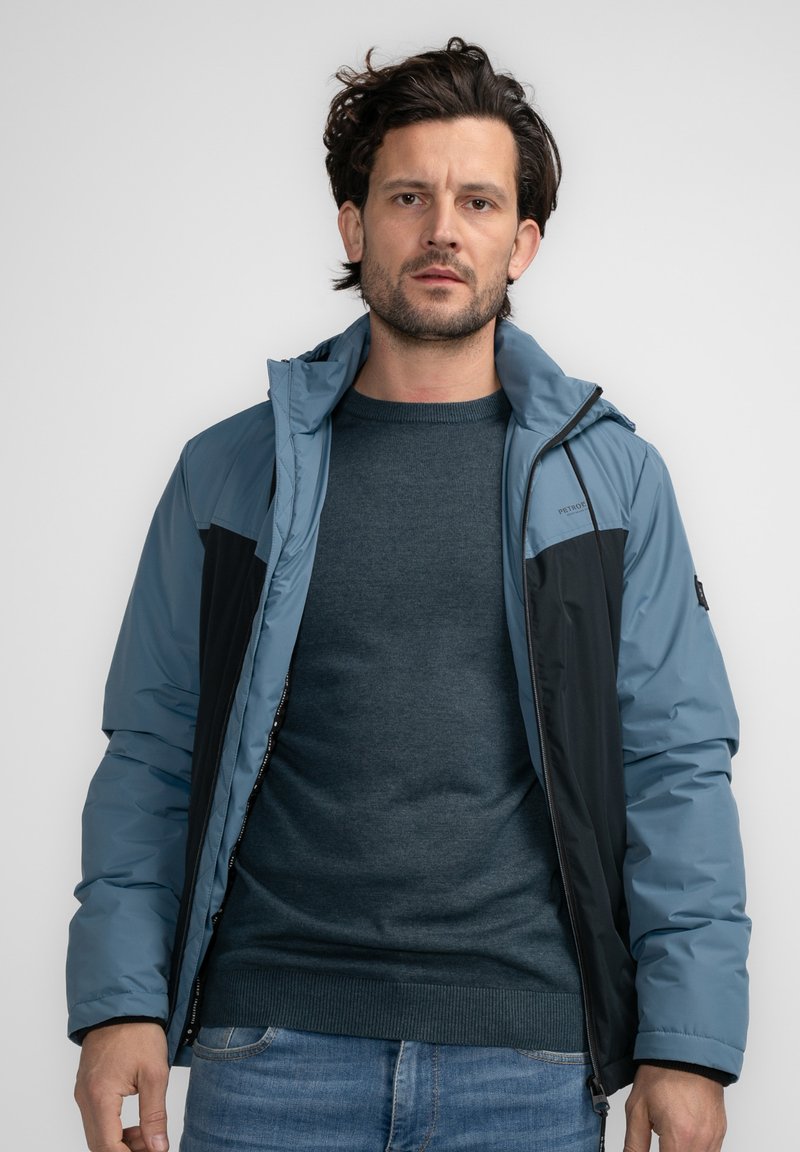 Chaleco aislante azul y negro con cierre de cremallera y capucha, que presenta una textura suave y paneles en contraste en los hombros.