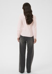 Femme aux longs cheveux bruns portant un t-shirt à manches longues rose clair et un jean large gris foncé, debout de dos sur un fond uni.