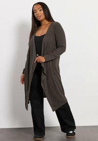 Lång, brun cardigan med öppna framsidor och en strukturerad mönstring, med långa ärmar och en fladdrande design. Kombinerad med svarta byxor och sneakers.