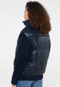Navy puffer vest with a glossy finish, high collar, and sleeveless design, worn over a textured navy sweater.