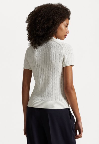 Žena s kudrnatými vlasy, oblečená v bílém krátkém rukávu svetr s copánkovým vzorem a tmavých kalhotách, zobrazena zezadu, stojící před jednoduchým pozadím.