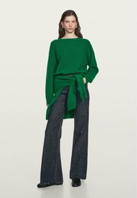 Maglione verde lavorato a maglia con vestibilità morbida, scollo ampio e maniche lunghe. Abbinato a jeans svasati scuri. Silhouette ampia e texture morbida.