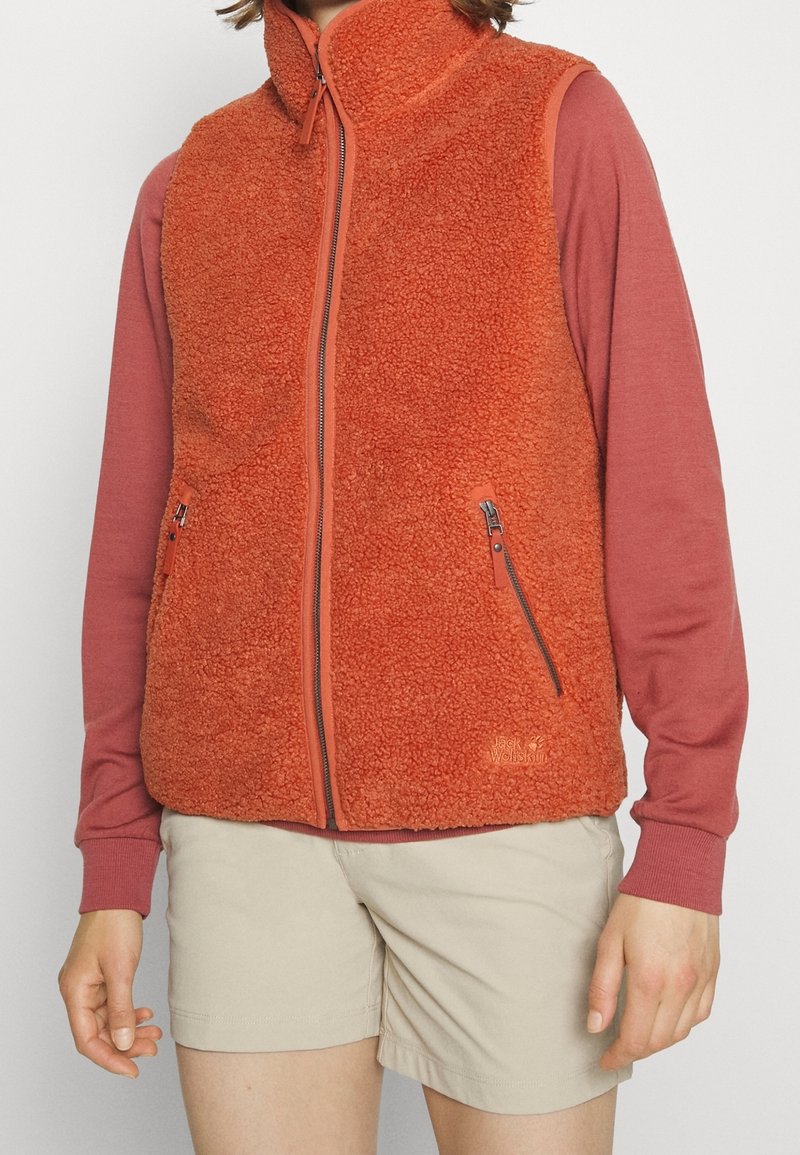 Gilet in pile arancione con colletto alto, zip frontale e due tasche laterali. Indossato sopra una maglietta a maniche lunghe rosa, abbinato a pantaloni corti beige.