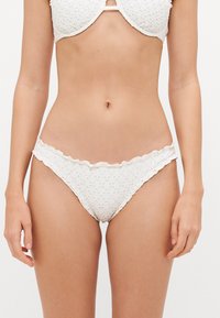 Bas de bikini blanc avec des bords festonnés, fabriqué à partir d'un tissu texturé présentant des motifs circulaires. La conception à l'avant et à l'arrière offre un ajustement couvrant complet.