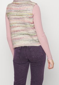 Gilet senza maniche lavorato a maglia con un motivo multicolore in morbidi toni pastello, indossato sopra una maglietta aderente rosa a maniche lunghe e abbinato a jeans viola.