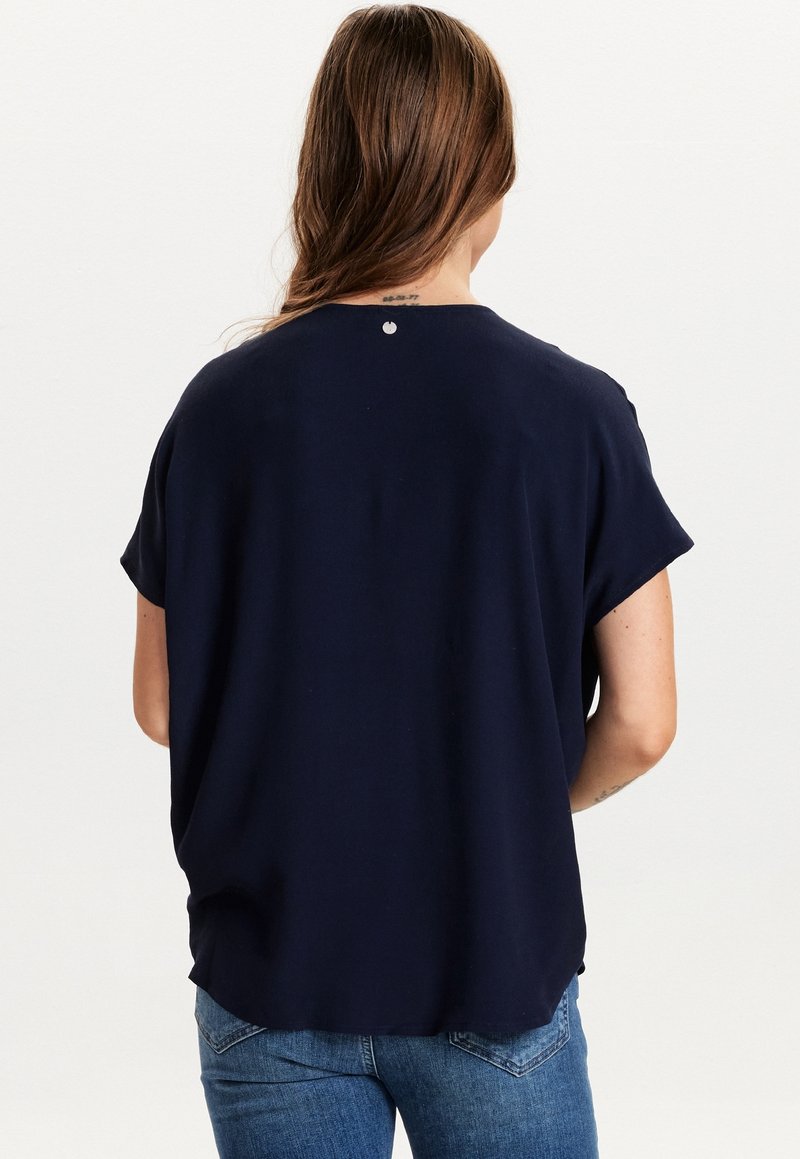 Granatowa bluzka z krótkim rękawem o luźnym kroju, z zaokrąglonym dołem i małym okrągłym akcentem na tyle przy dekolcie. Gładka faktura materiału.