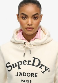 Crème-kleurige hoodie met een trekkoordkap, voorzien van een zwart gedrukt logo, gedragen over een lichtroze gestreept overhemd met kraag.