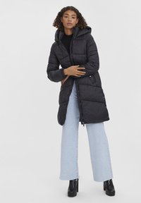Doudoune noire avec capuche, fermeture éclair à l'avant, poches latérales et texture matelassée. Portée sur un jean large bleu clair et des bottes à plateforme noires.