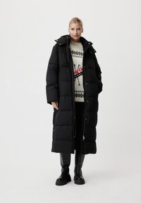 Femme portant un long manteau en duvet noir, des bottes noires et un pull blanc avec un motif noir et rouge, se tenant devant un fond clair et uni.