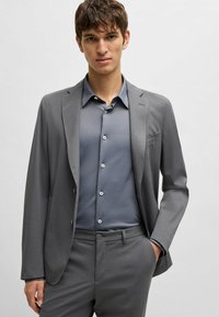 Grau Anzugjacke und Hose kombiniert mit einem grauen Hemd mit Knöpfen. Der Stoff wirkt glatt. Der Anzug hat geknöpfte Revers und zwei Knöpfe.