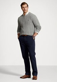 Moški, ki nosi sivega pulover z vzorcem pletenice preko bele srajce, mornarsko modre hlače, rjav pas, rjave usnjene čevlje in ročno uro, stoji z rokami v žepih.