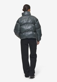 Dik donkergrijs jasje met hoge kraag en horizontale gestikte secties, gecombineerd met zwarte broek. Glad, glanzend en geïsoleerd materiaal.
