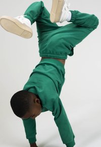 Groene katoenen sweatshirt en broek gedragen door een kind dat een handstand doet, met witte sneakers met beige zolen, tegen een effen achtergrond.