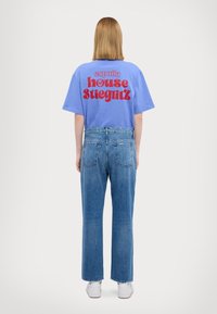 Vaaleansininen lyhythihainen t-paita, jossa on punaista tekstiä selkäpuolella, yhdistettynä suoriin sinisiin farkkuihin ja valkoisiin tennareihin.