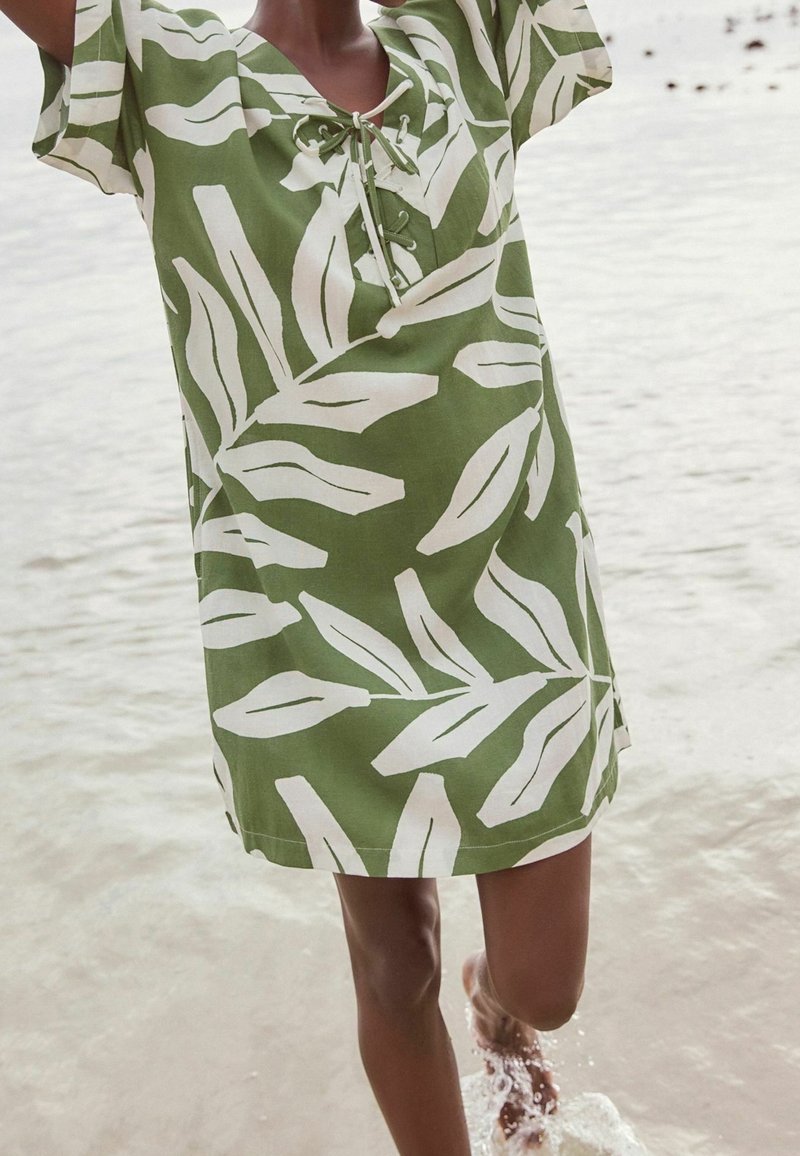 Personne portant une robe verte à manches courtes avec un motif de feuilles blanches, debout dans une eau peu profonde en éclaboussant avec un pied.