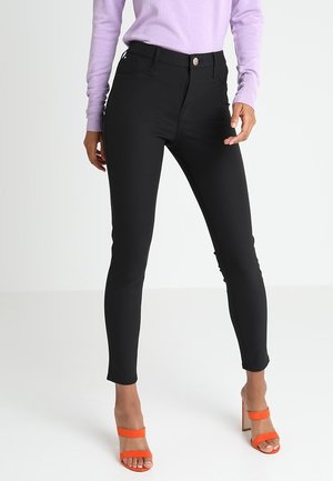 Zwarte skinny jeans met een hoge taille, voorzien van zijzakken en een gladde stofstructuur. Het model combineert ze met oranje sandaaltjes met bandjes.