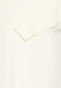 Chemise à manches longues en tissu blanc doux. Elle présente un poignet légèrement froncé et une texture lisse, avec une coupe décontractée.