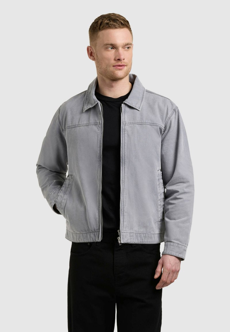 Homme portant une veste zippée gris clair par-dessus un t-shirt noir et un pantalon noir, debout avec une main dans la poche, regardant sur le côté.