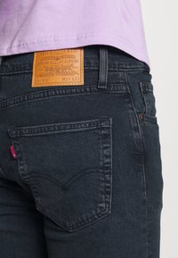 Zadní pohled na tmavě modré džíny Levi's s koženou nášivkou značky a osobu oblečenou v světle fialové košili, která je mírně zasunuta nad pas.