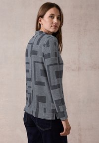 Maglietta grigia a maniche lunghe con un motivo geometrico di linee bianche. Tessuto morbido, scollatura rotonda e taglio dritto nell'orlo. Indossata con jeans scuri.