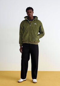Zaļa kordūrā zip-hoodie ar priekšējām kabatām, ribotām piedurknēm un apakšmalu; saskaņota ar melniem plats kāju biksēm un baltām kurpēm.