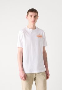 Maglietta bianca in cotone con un piccolo logo arancione sul lato sinistro del petto. Collo rotondo, maniche corte e vestibilità comoda. Abbinata a pantaloni beige.