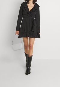 Vestido negro envolvente con escote en V, detalles fruncidos, mangas largas y dobladillo acampanado. Combinado con botas vaqueras negras y un bolso gris.