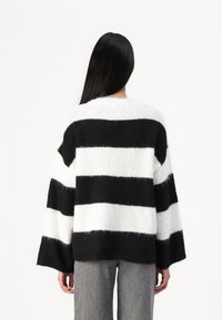 Personne aux longs cheveux noirs portant un pull ample à larges rayures noires et blanches et un pantalon gris, vue de dos sur un fond blanc.
